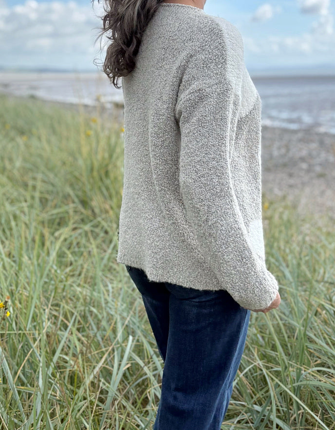 Person wearing a patterned boucle sweater standing in a grassy field with a blue sky and water in the background