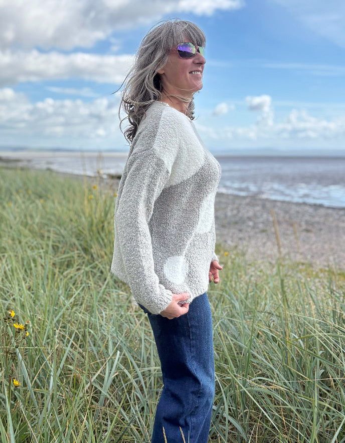 Person wearing a patterned boucle sweater standing in a grassy field with a blue sky and water in the background