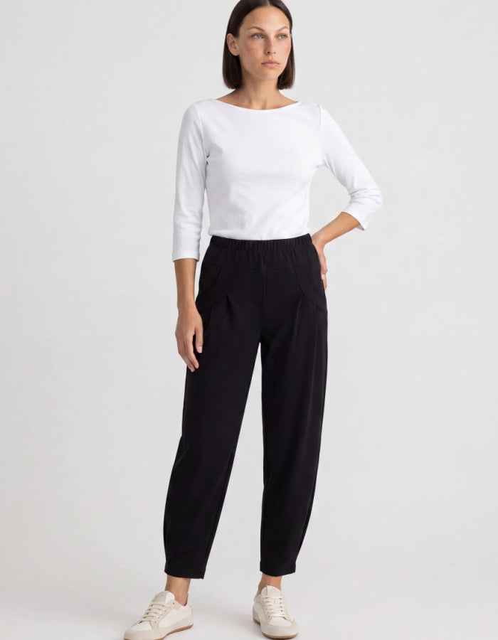 A woman modeling a white long-sleeve top and black wide-leg cropped pants with white sneakers, standing against a light gray studio background.