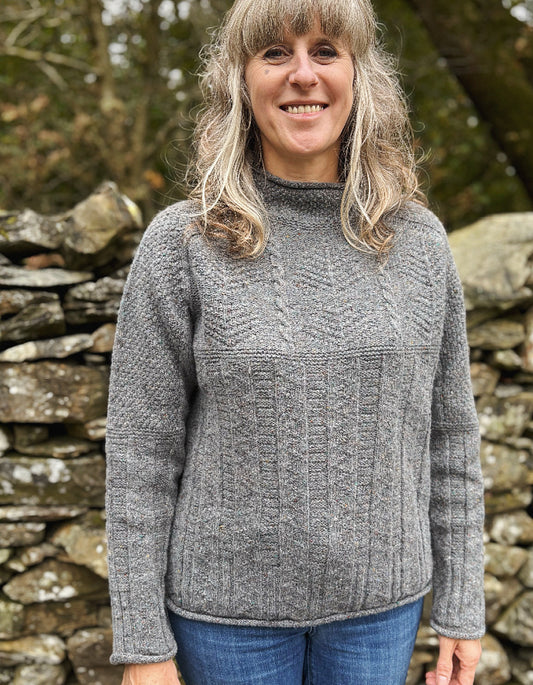 Woman wearing a gray cable knit sweater standing in front of a stone wall.
