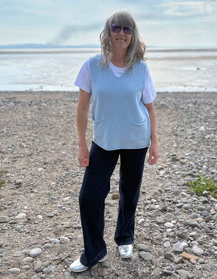 Person standing on a pebbly beach wearing a light blue top and dark wide leg jeans
