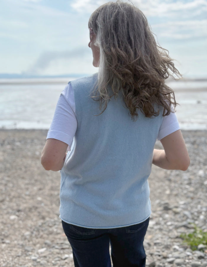 Person standing on a pebbly beach looking out towards the ocean. light blue wool sweater vest