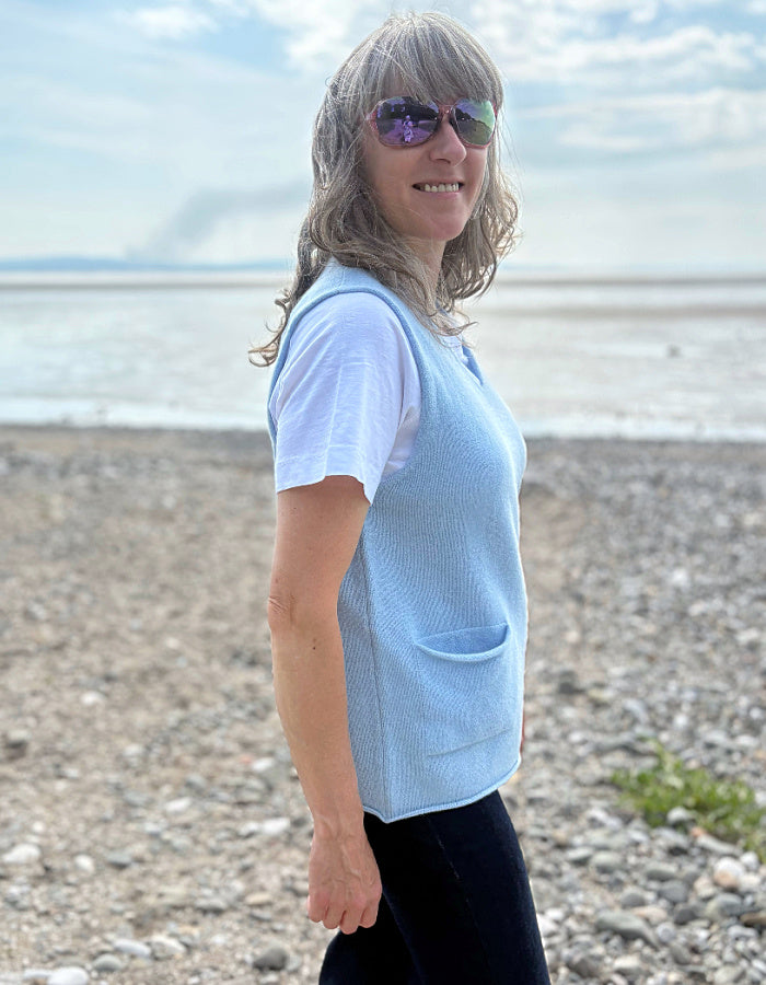 Woman wearing a light blue vest over a white shirt on a pebbly beach.