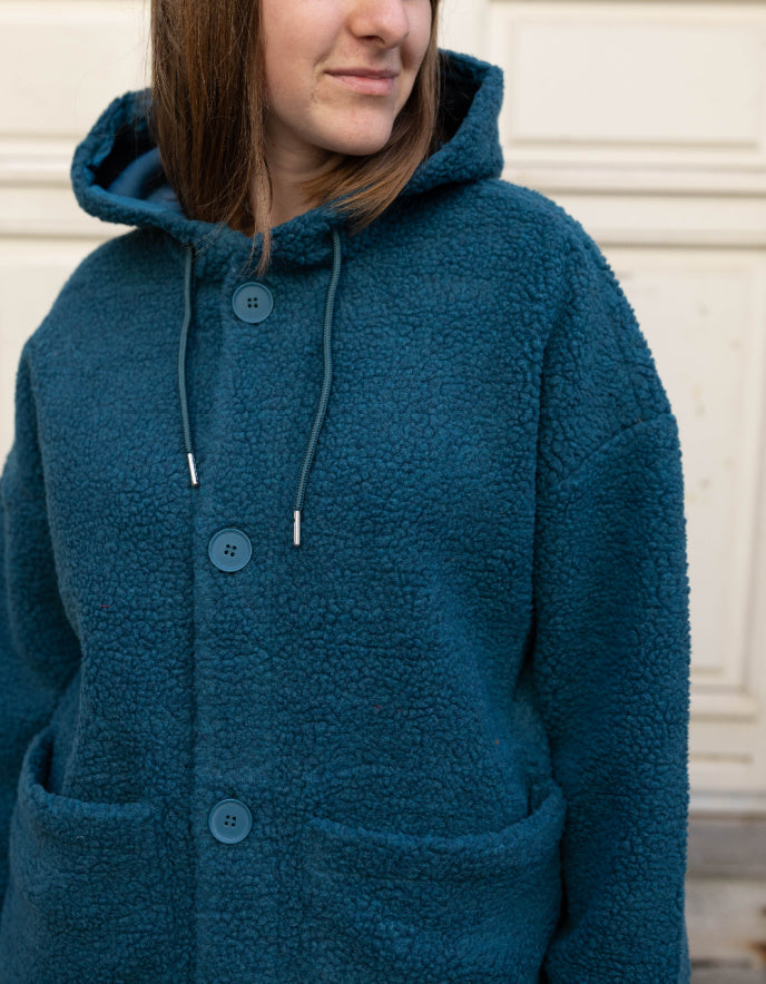 Person wearing a blue hooded coat with buttons against a neutral background