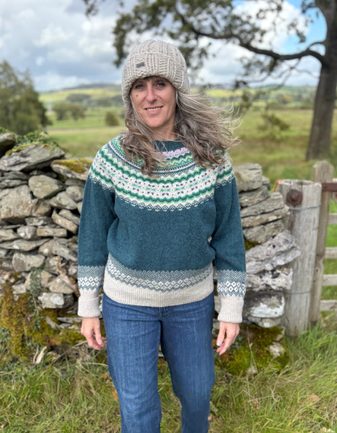 Woman wearing a patterned sweater and knit hat standing in a rural landscape with stone walls and green fields.