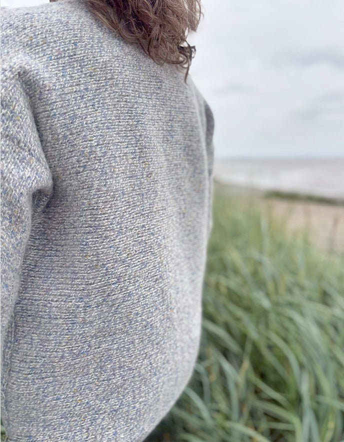 Person wearing a silver grey donegal tweed sweater and sunglasses standing in a grassy field with a cloudy sky.