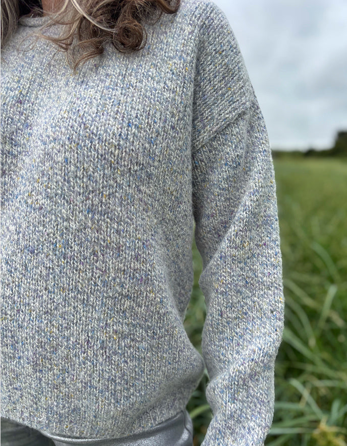 Person wearing a silver grey donegal tweed sweater and sunglasses standing in a grassy field with a cloudy sky.