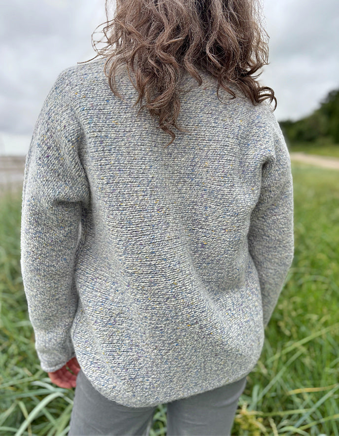 Person wearing a silver grey donegal tweed sweater and sunglasses standing in a grassy field with a cloudy sky.