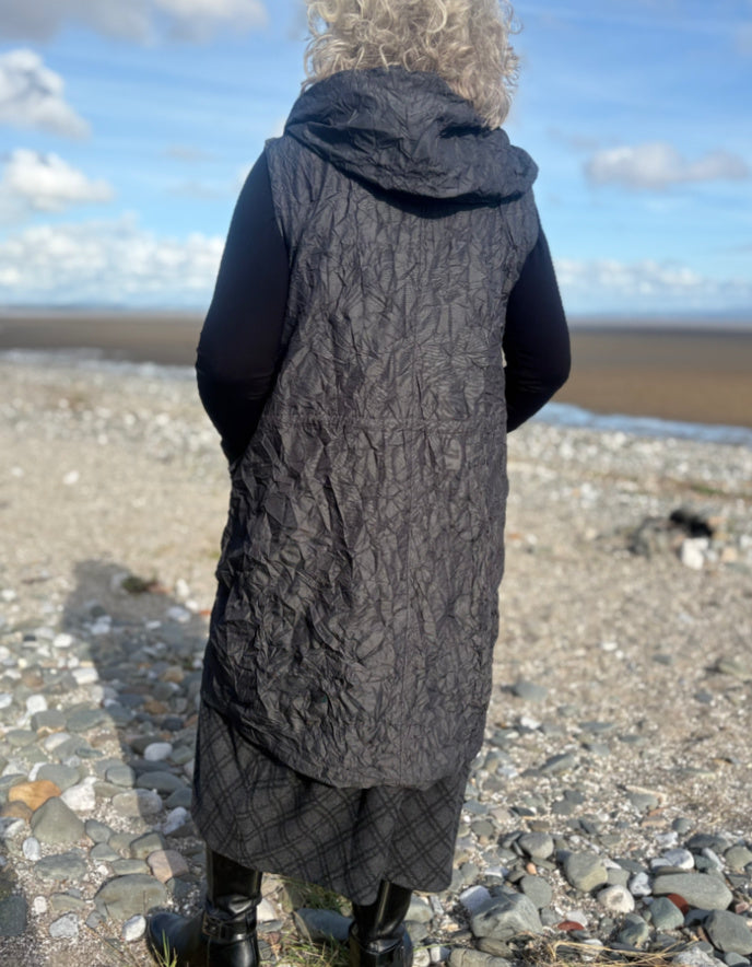 Person wearing a long dark gray quilted vest standing on a pebbly beach with a blue sky.