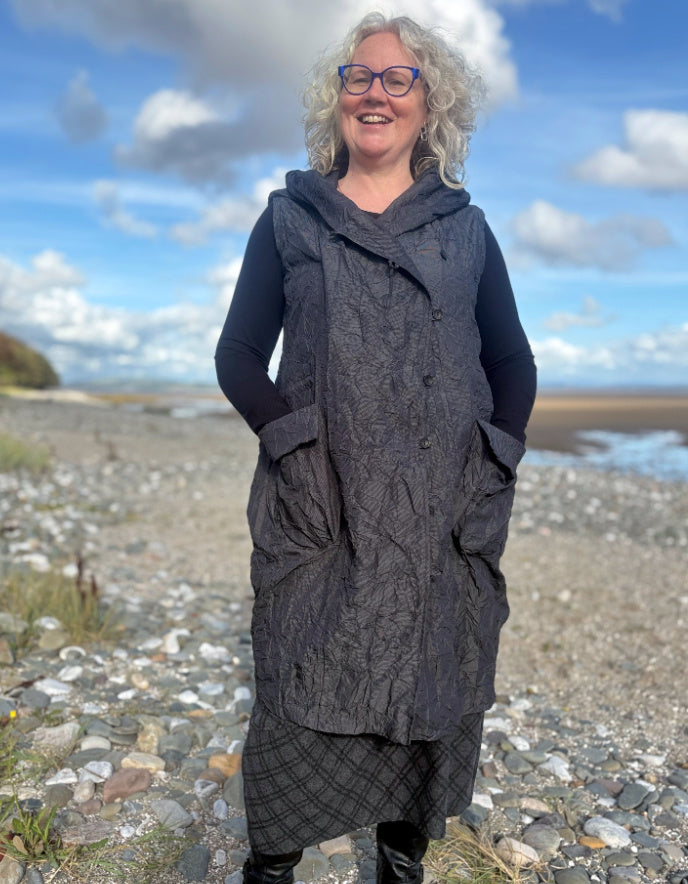 Woman wearing a long dark gray knit vest on a beach with a blue sky and clouds in the background.