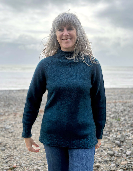 Woman wearing a teal sweater standing on a pebbly beach with ocean and cloudy sky in the background