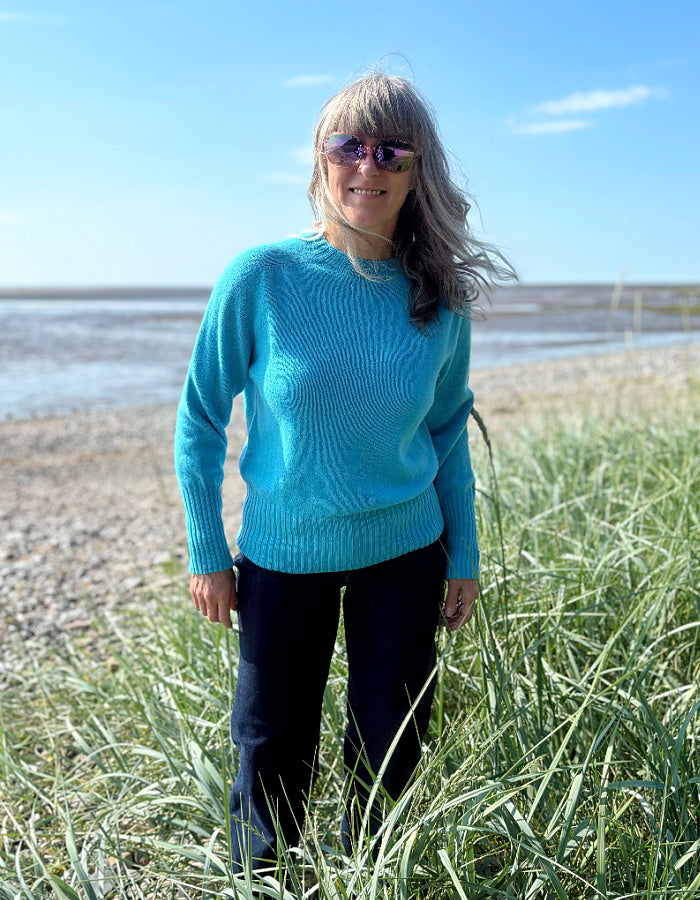 Person wearing a wool blue turquoise sweater standing in tall grass by a beach.