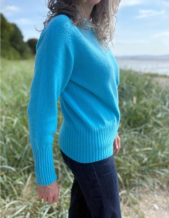 Person wearing a wool blue turquoise sweater standing in tall grass by a beach.