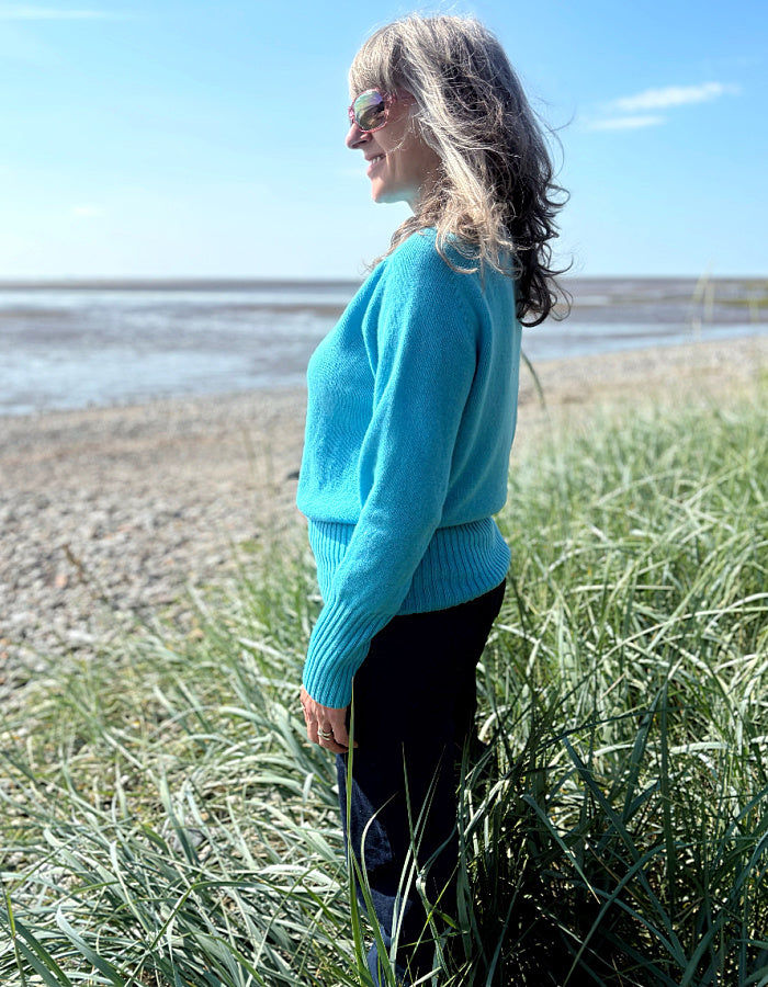 Person wearing a wool blue turquoise sweater standing in tall grass by a beach.