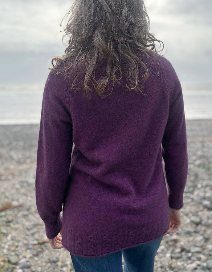 Person wearing a purple sweater standing on a pebbly beach with a cloudy sky.