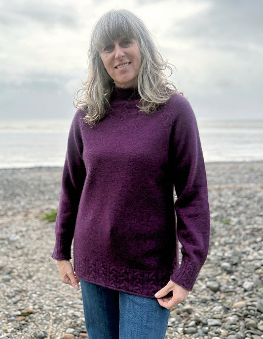 Woman wearing a purple sweater standing on a pebbly beach with water in the background