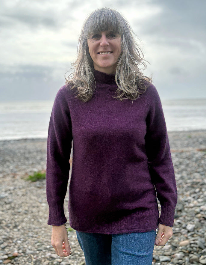 Woman wearing a purple sweater standing on a pebbly beach with water and sky in the background