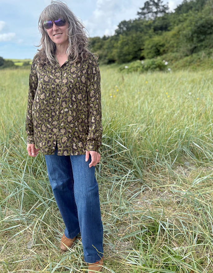 Woman wearing a leopard print top and blue jeans standing in a grassy field with trees in the background.