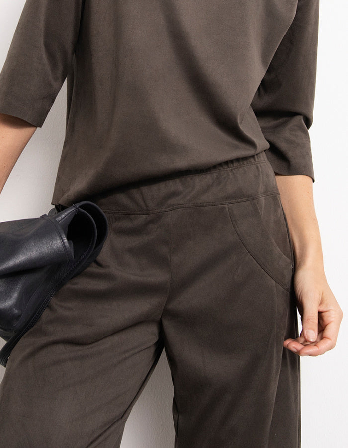 Close-up of a person wearing darl olive wide-leg trousers and a matching dark brown top, holding a black leather clutch against a plain light background.