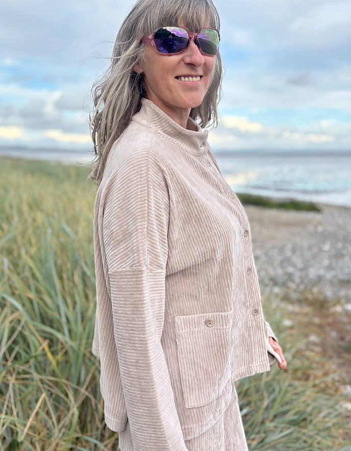 Person wearing a winter white jumbo cord jacket and sunglasses on a beach with grass and water in the background