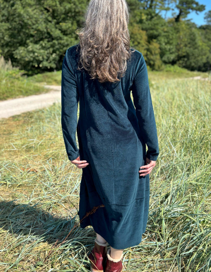 Woman in a long dark teal velvet dress with long sleeves and V neck standing in a field with trees in the background