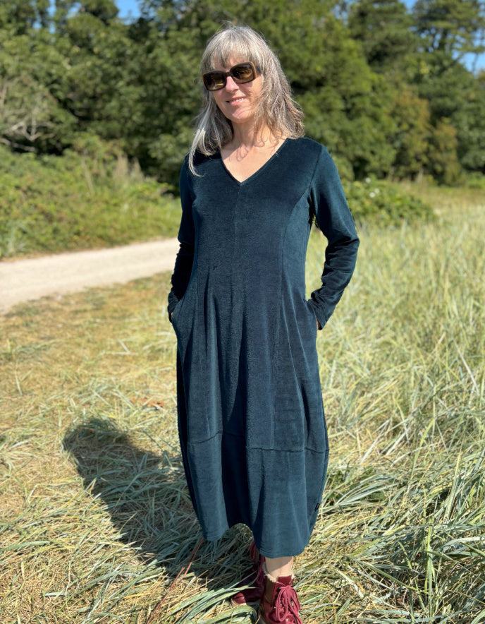 Woman in a long dark teal velvet dress with long sleeves and V neck standing in a field with trees in the background