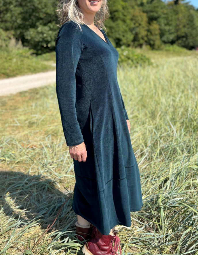 Woman in a long dark teal velvet dress with long sleeves and V neck standing in a field with trees in the background
