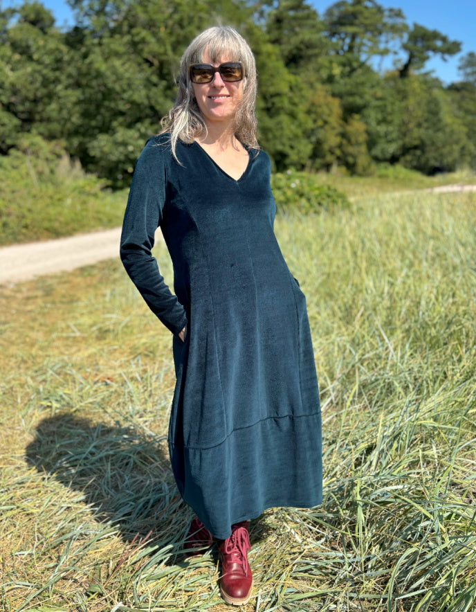 Woman in a long dark teal velvet dress with long sleeves and V neck standing in a field with trees in the background