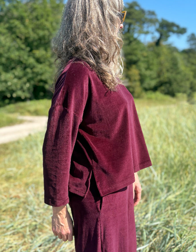 Person wearing a burgundy velvet top and trouser outfit standing in a field with trees in the background