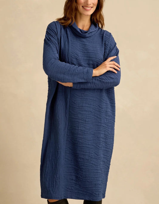 A woman wearing a textured, long-sleeve cowl-neck dress in a dusty navy blue, standing with her arms crossed against a solid beige background.