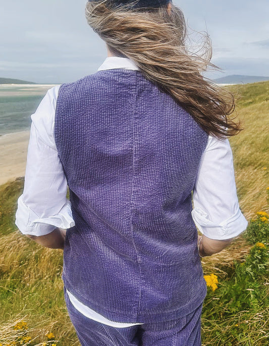 A woman viewed from behind wearing a textured purple corduroy vest over a white shirt, standing on a grassy coastal dune with the sea and cloudy sky in the background.