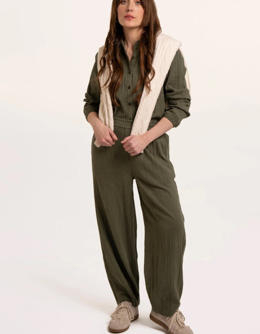 A woman modelling an olive green checked barrel leg trousers with a cream ribbed knit vest layered over it, paired with beige sneakers, against a plain white studio background.