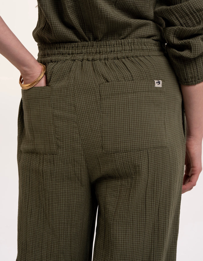 A woman modelling an olive green checked barrel leg trousers paired with beige sneakers, against a plain white studio background.