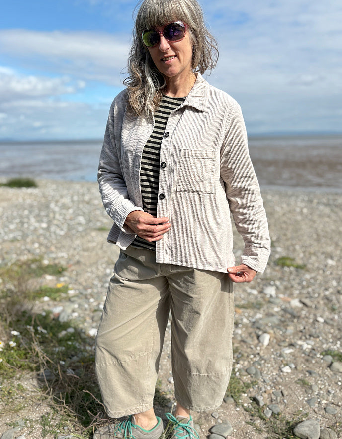 Woman standing on a pebbly beach wearing sunglasses and cream cord shirt, stripes t shirt and beige cord lantern trousers