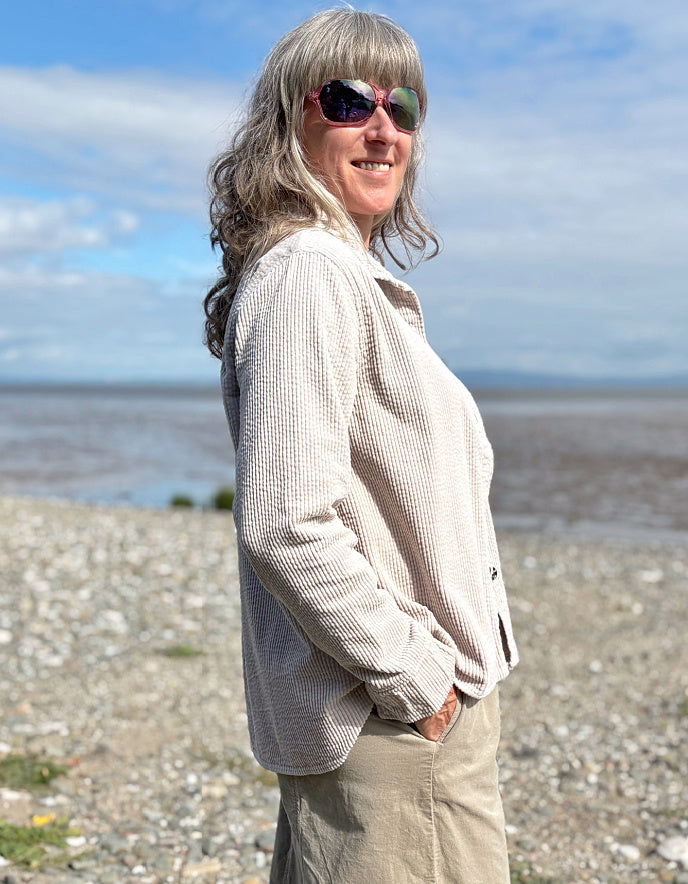 Woman standing on a pebbly beach wearing sunglasses and cream cord shirt, stripes t shirt and beige cord lantern trousers