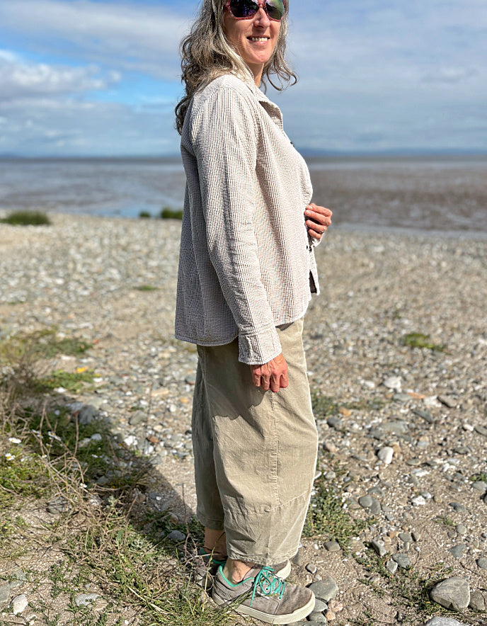 Woman standing on a pebbly beach wearing sunglasses and cream cord shirt, stripes t shirt and beige cord lantern trousers