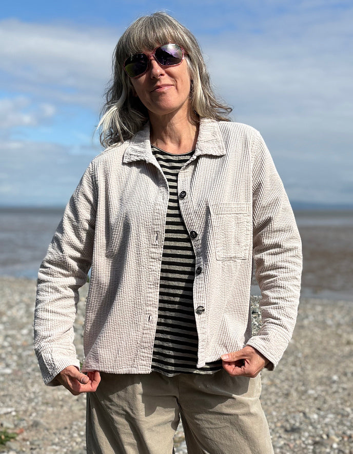 Woman standing on a pebbly beach wearing sunglasses and cream cord shirt, stripes t shirt and beige cord lantern trousers