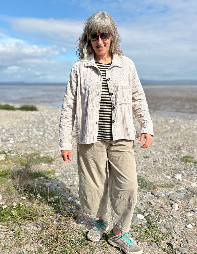 Woman standing on a pebbly beach wearing sunglasses and cream cord shirt, stripes t shirt and beige cord lantern trousers
