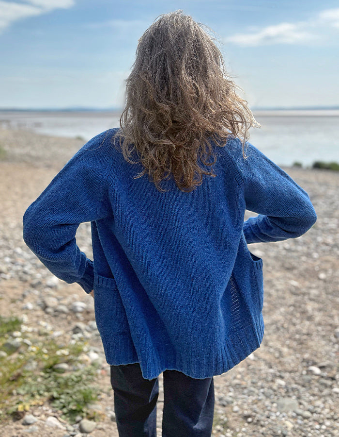 Woman wearing a wool blue cardigan on a pebbly beach with water and sky in the background