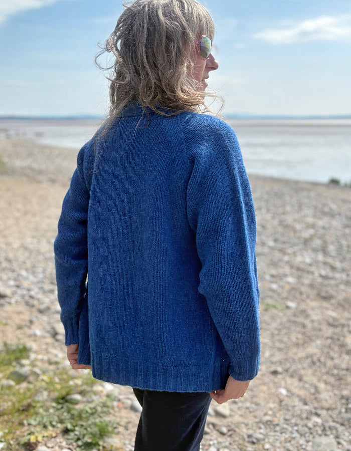 Woman wearing a wool blue cardigan on a pebbly beach with water and sky in the background