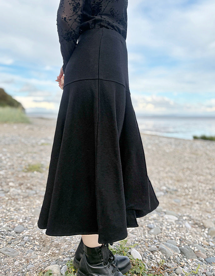 Person wearing a black lace top and black wool skirt standing on a pebbly beach with ocean and sky in the background.