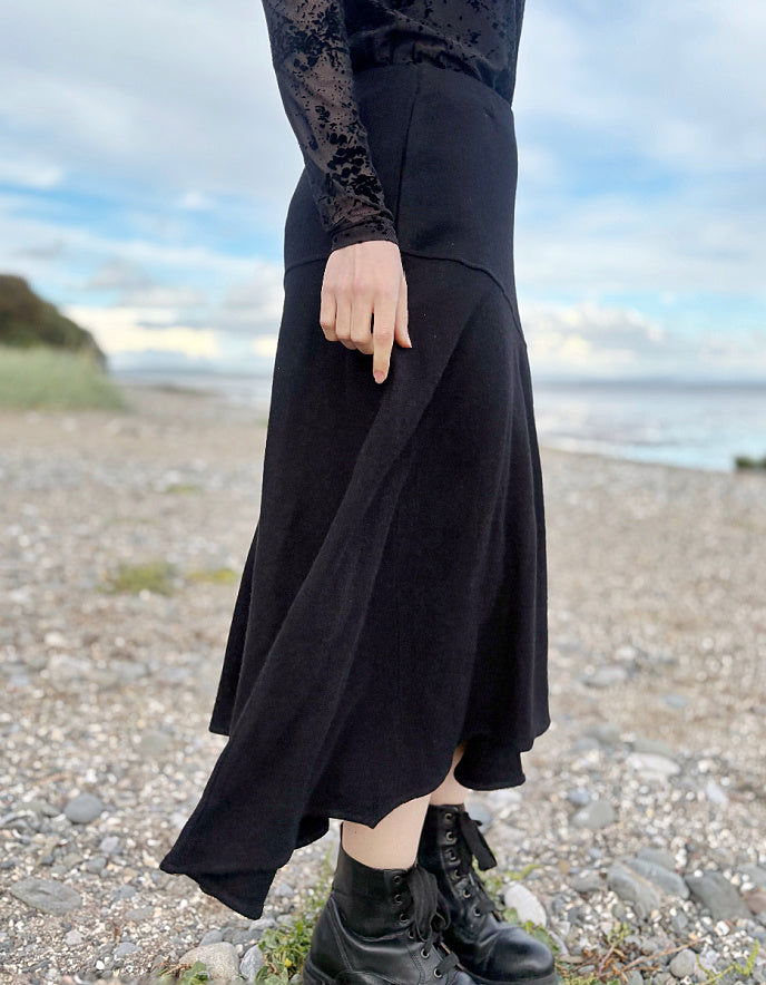 Person wearing a black lace top and black wool skirt standing on a pebbly beach with ocean and sky in the background.
