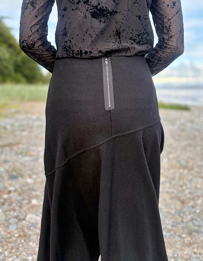 Person wearing a black lace top and black wool skirt standing on a pebbly beach with ocean and sky in the background.