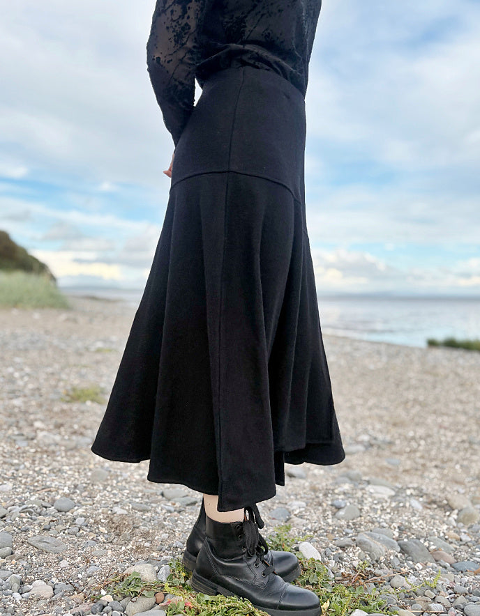 Person wearing a black lace top and black wool skirt standing on a pebbly beach with ocean and sky in the background.