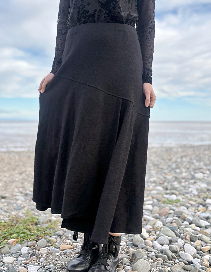 Person wearing a black lace top and black wool skirt standing on a pebbly beach with ocean and sky in the background.