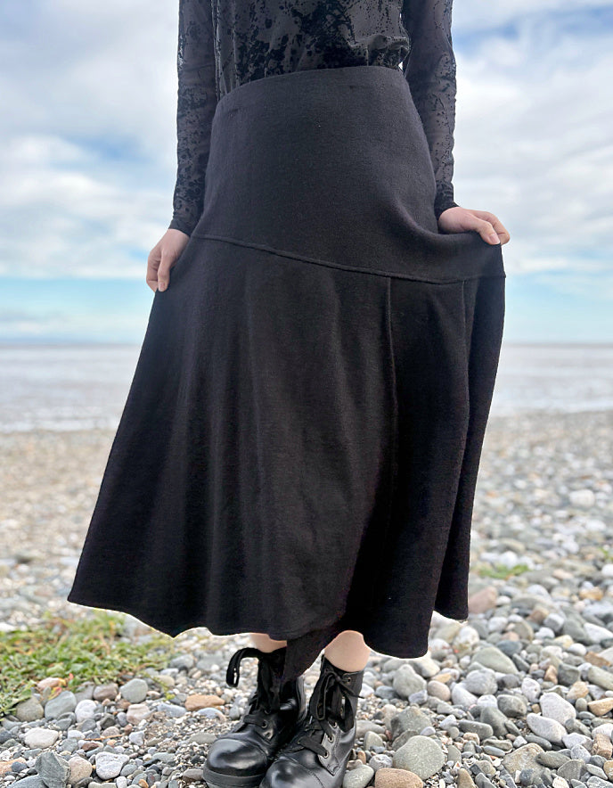 Person wearing a black lace top and black wool skirt standing on a pebbly beach with ocean and sky in the background.