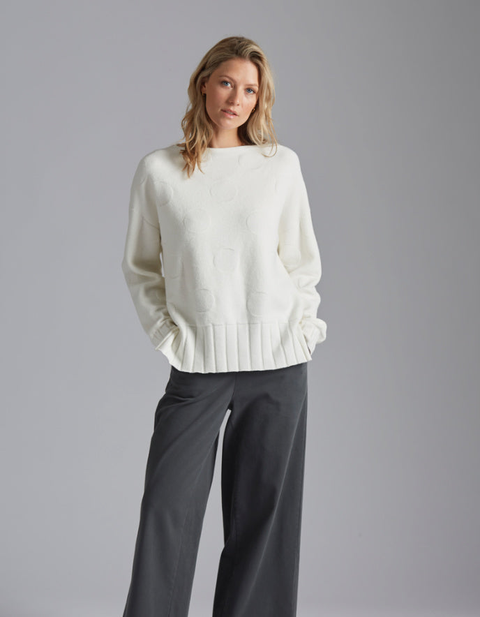 Woman wearing a white sweater and dark pants against a gray background