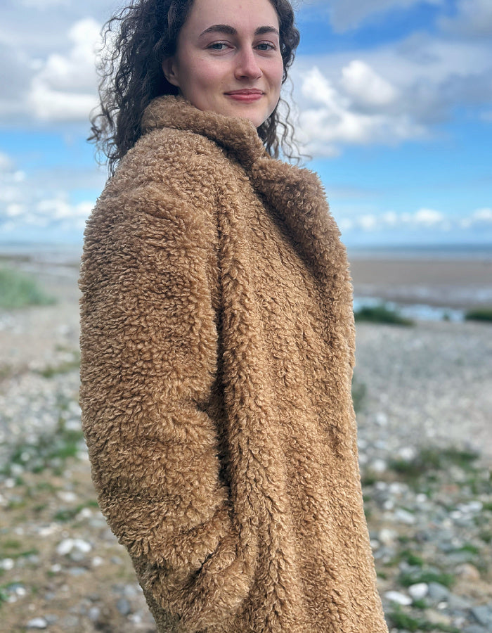A woman wearing a warm camel brown sherpa fleece jacket, photographed outdoors on a pebble beach with a cloudy blue sky in the background.