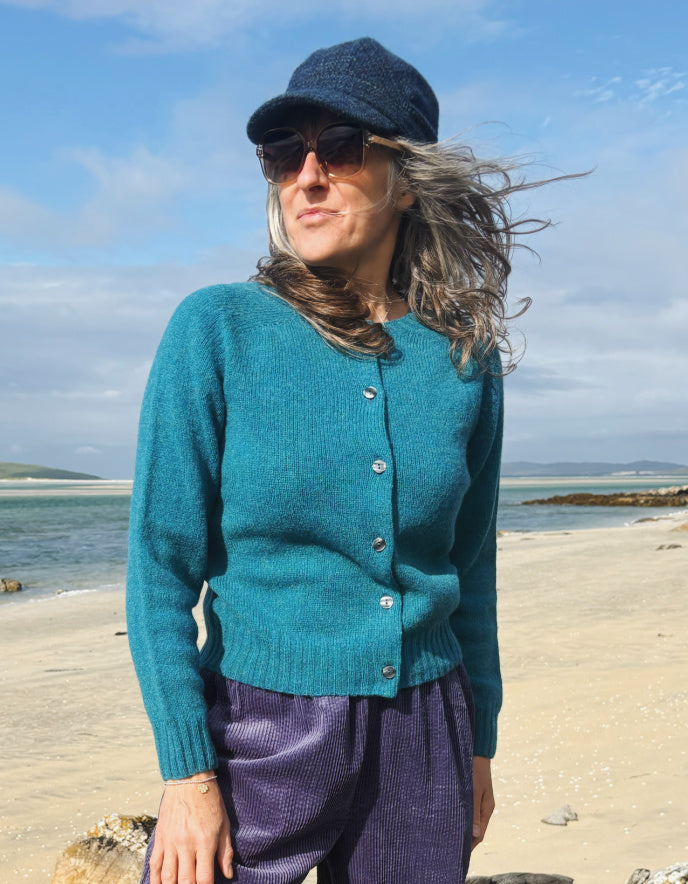 A woman standing on rocks by a beach wearing a teal button-front cardigan, purple corduroy pants, and white sneakers.