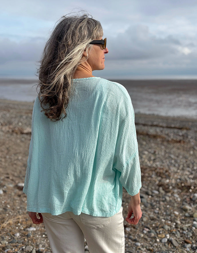 Woman in a light blue shirt and sunglasses standing on a pebbly beach.